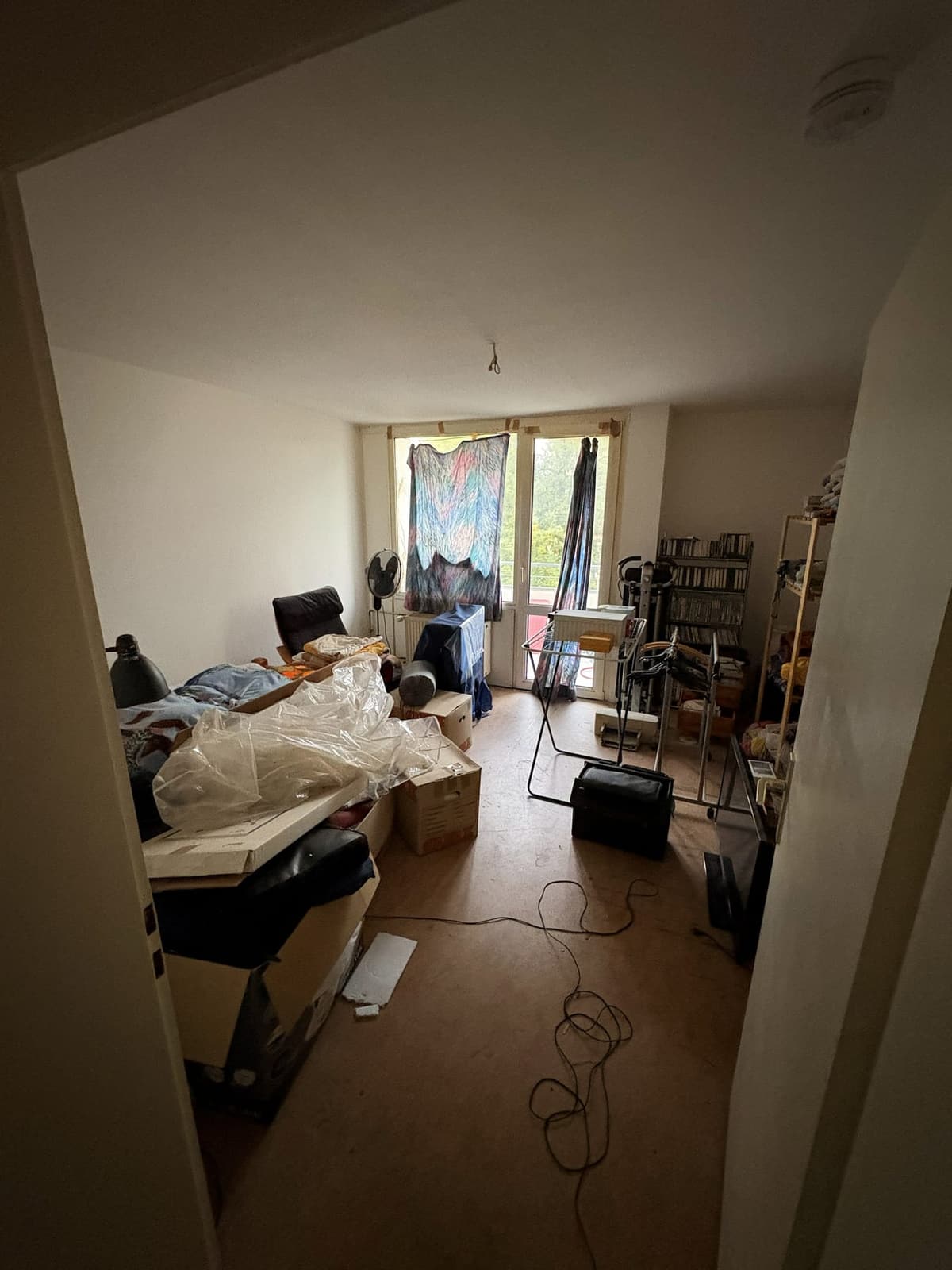 Zimmer mit Balkontür vor der Wohnungsauflösung, vom Flur aus gesehen, vollgestellt mit Möbeln und Kartons